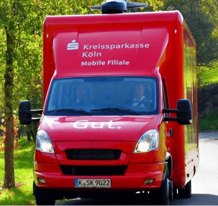 Kraftfahrzeug der Kreissparkasse Köln, roter Kreditservice-Fahrzeug mit der Aufschrift "Gut", fährt im Freien durch eine grüne, baumbestandene Umgebung.