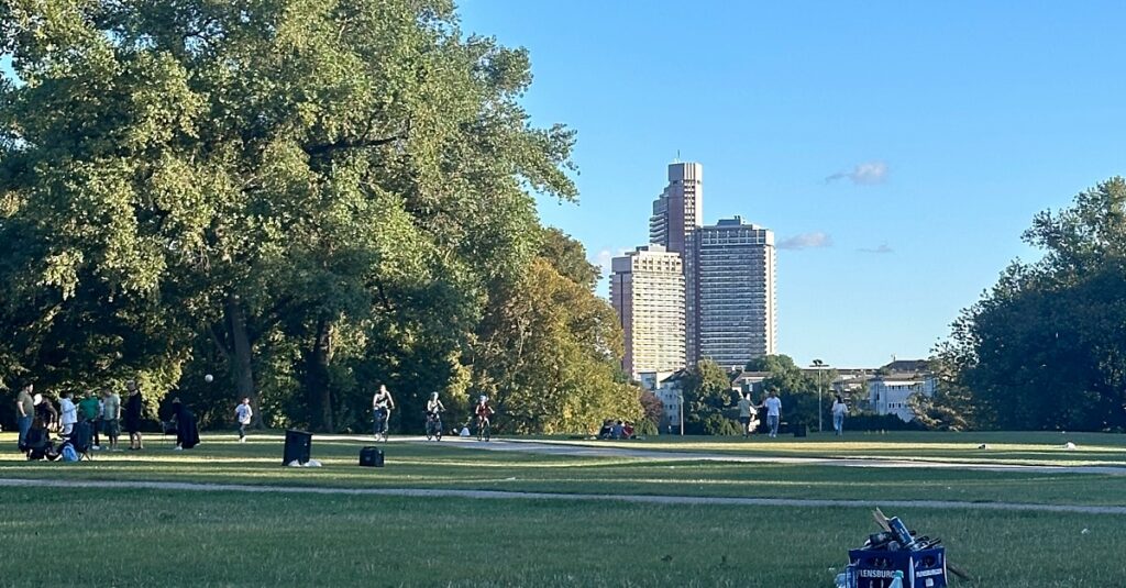 Grüner Stadtpark mit hohen Bäumen und Blick auf moderne Hochhäuser in der Stadtmitte, ideal zum Entspannen und Spazierengehen.