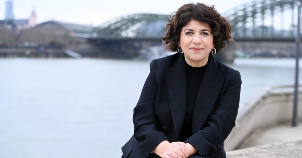 Lindweiler Frau am Flussufer in einer schwarzen Blazer, mit Brücke im Hintergrund, urbane Szenerie, entspannte Atmosphäre, professionelle Ausstrahlung, modern gekleidet, stilvoll, Business-Umfeld, städtischer Kontext, elegant.