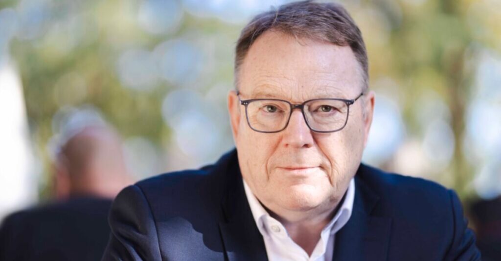 Mann in formellem Outfit mit Brille, Blick direkt in die Kamera, im Freien bei Tageslicht, unscharfer Hintergrund, professionelle Portraitaufnahme.