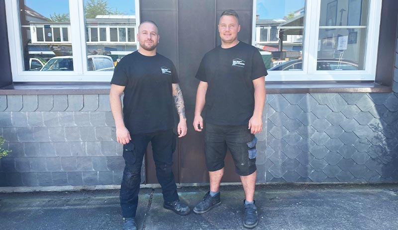 Zwei Männer in schwarzen T-Shirts stehen vor einem Gebäude mit dunkler Fassade und großen Fenstern, repräsentieren das Team von Lindweiler.