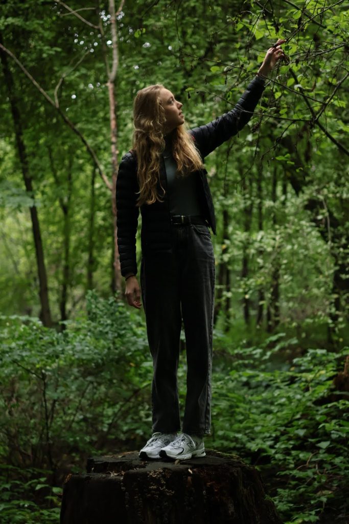 Helkroppsporträtt på en ung tjej som står på en stor brun stubbe ute i skogen. Runt henne är det lysande grön natur och hon sträcker sin vänstra hand efter ett träds gren. Hennes blick möter hennes hand vis grenen hon sträcker sig efter.