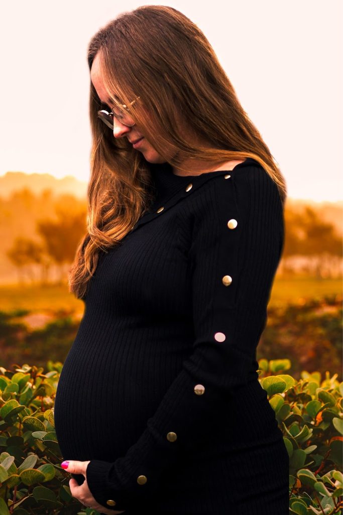 Porträtt på en gravid kvinna. Bilden är fotograferad från sidan där hon tittar ned på hennes mage som hon håller under med sin vänstra hans. Hon tittar med längtan i blicken. Bakgrunden är ljus och liknar Golden Hour.