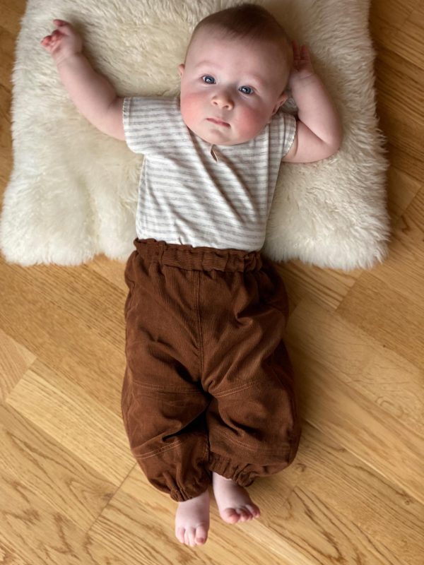 baby der ligger på en pelspude poå gulvet i brune bukser og hvid stribet top