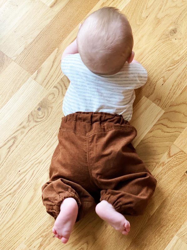 Baby der ligger på et trægulv i brune fløjsbukser og hvid stribet bluse