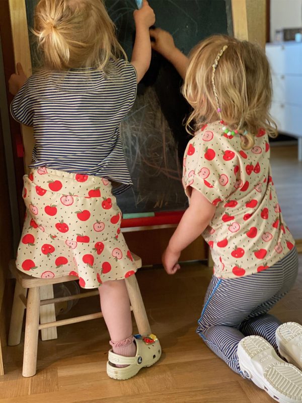 2 girls drawing on a blackboard in clothes with blue stripes and red apples printed on blouse and skirt