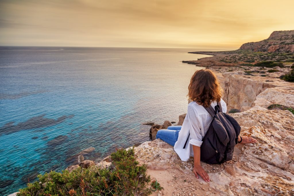 A stylish young woman traveler watches a beautiful sunset on the rocks on the beach, Cyprus, Cape Greco, a popular destination for summer travel in Europe