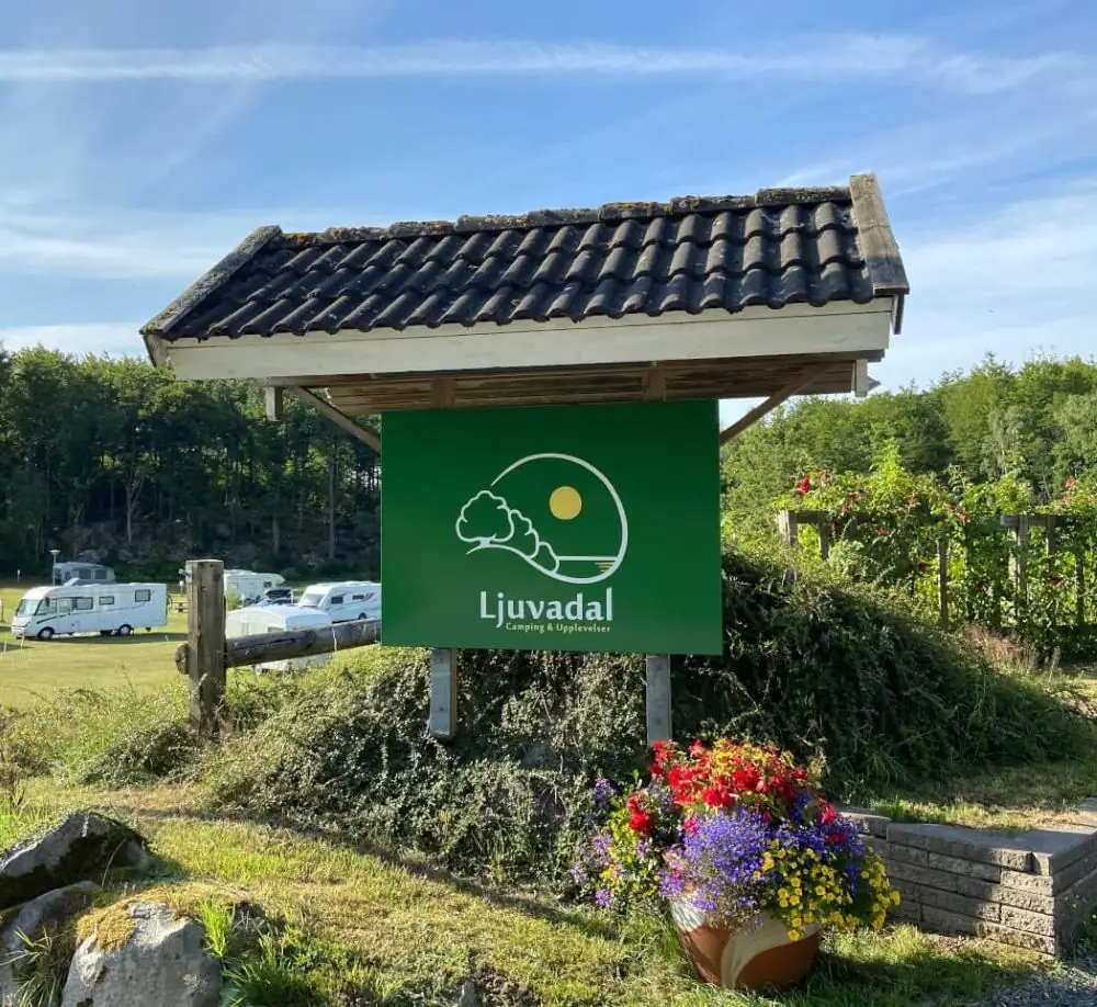 Grünes Eingangsschild mit dem Logo von Ljuvadal Camping & Upplevelser, umgeben von einem Fahnenmast und Blumen.