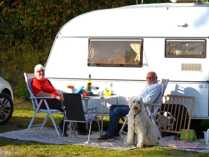 Ein Mann und eine Frau sitzen mit einem weißen Hund vor ihrem Wohnwagen auf Ljuvadal, ein Camping in Blekinge.