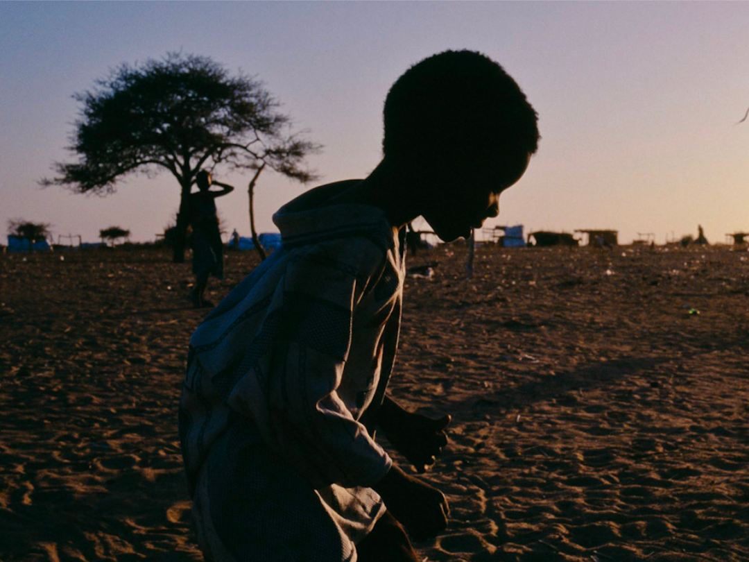 Boy Silhouette of a child crosses in front of the camera with a silhouette of a woman carrying a bundle on her head in the background (Darfur, Sudan)