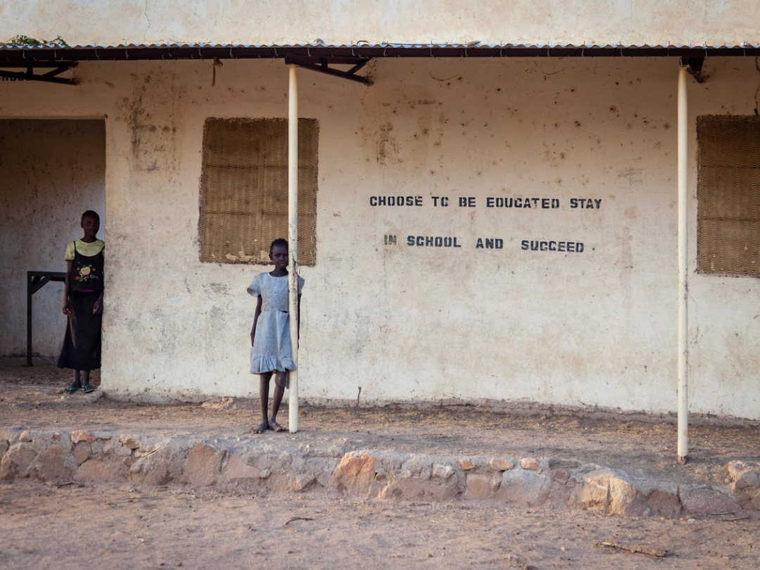 Choose to be educated Two girls stand in front of a building looking toward the camera, one leans against a metal support the other in the doorway. The side of the building reads Choose to be educated stay in school and succeed