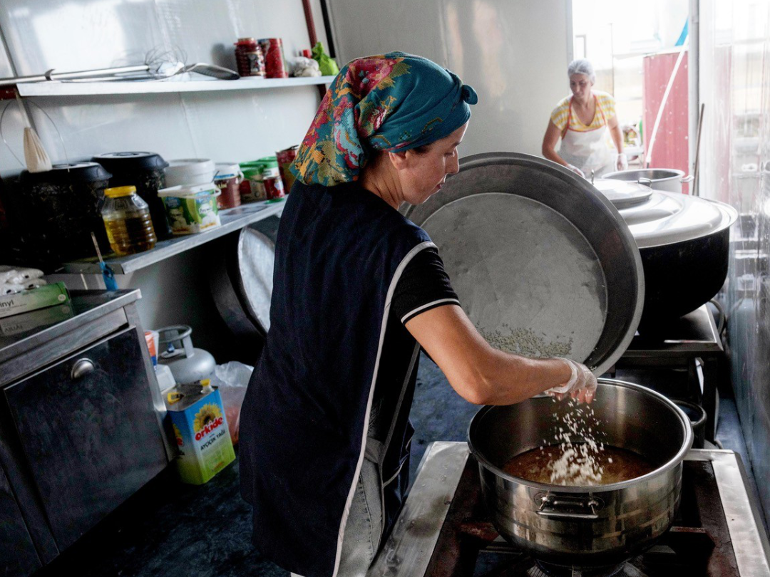 Cooking meals for many A woman in a blue headscarf and apron pours rice from a large dish into a saucepan above a gas hob