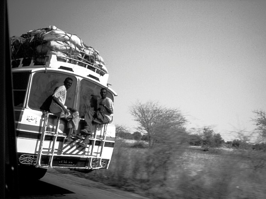 riding the bus Black and white photo of two men sitting on the metal frame on the back of a bus loaded with bags on top.