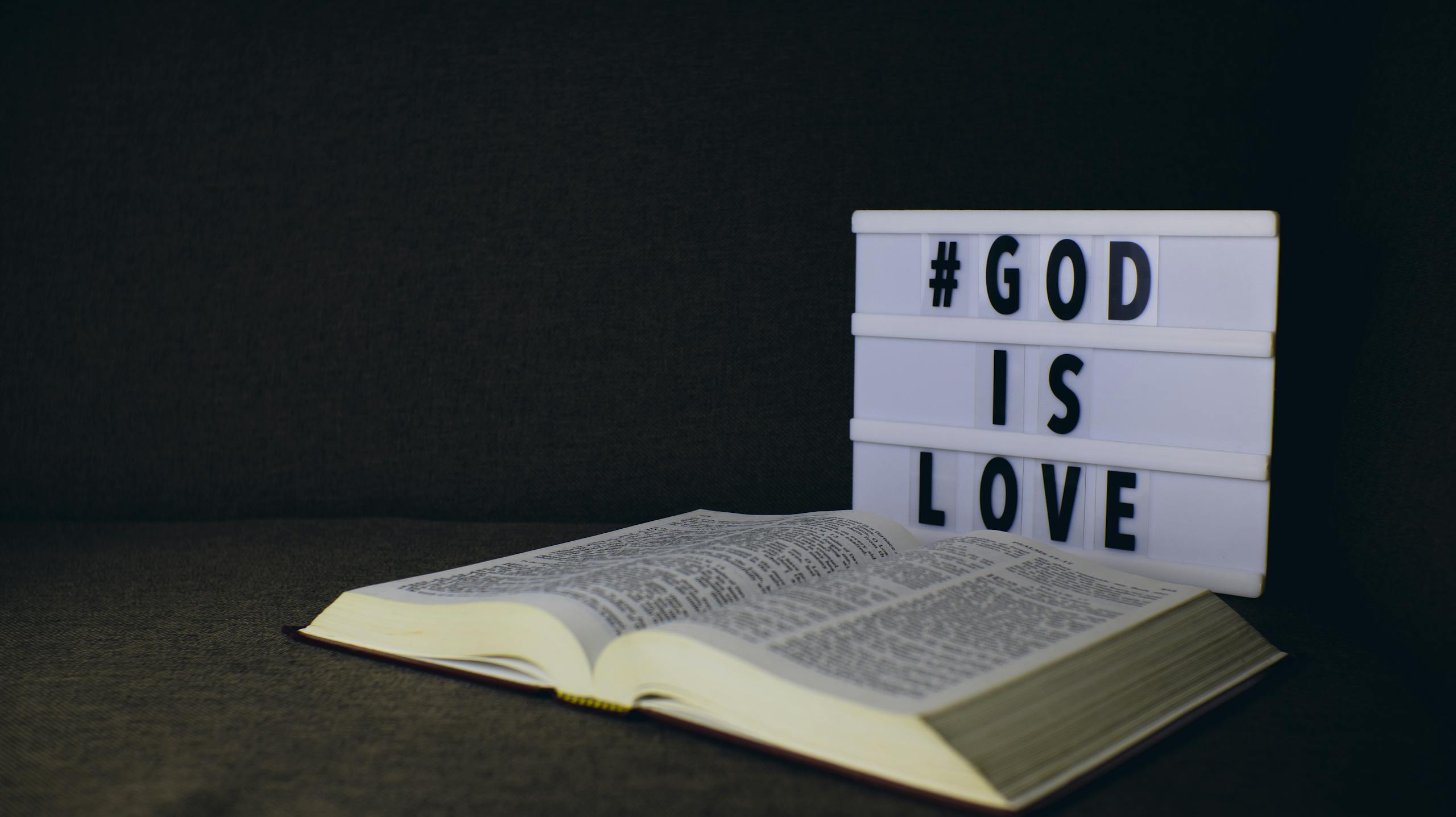 An open Bible next to a lightbox sign displaying the text 'God is Love' on a dark background.