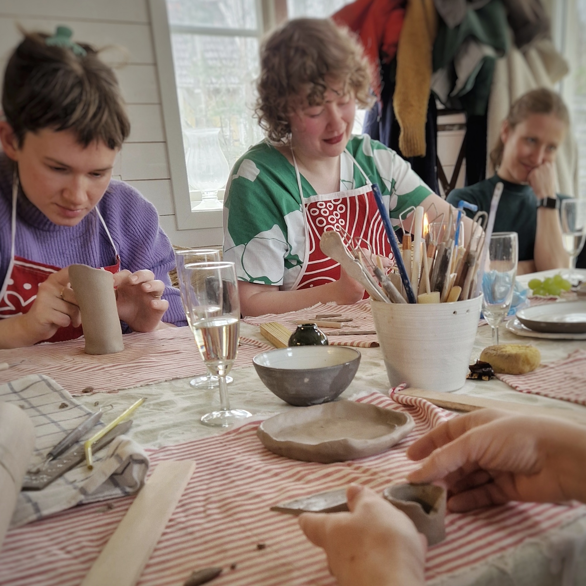 tre unga kvinnor skapar i lera i en kreativ keramikstudio. På bordet syns pågående skapande och bubbel i glas