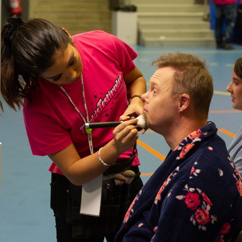 Manlig artist sminkas av sminkös innan uppträdande på Funkisfestival.