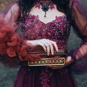 Nainen purppuran värisessä juhlavassa mekossa pitää koristeellista rasiaa, jonka raosta nousee punaista savua, käsissään