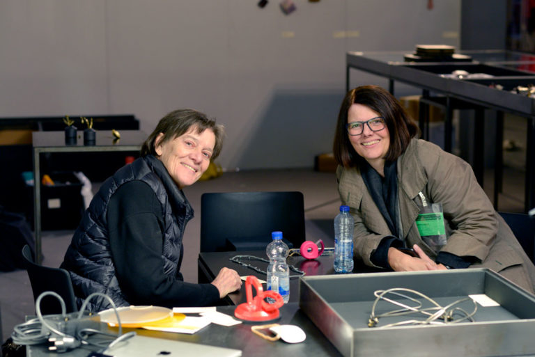 Marie-José van den Hout and Petra Hoelscher at FRAME 2019 during the building up of the stand of Galerie Marzee - on the table a necklace by Lucy Sarneel and a necklace by Dorothea Prühl