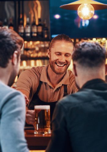 Cheerful bartender serving beer to young men while standing at the bar counter in pub