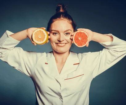 A lady in a lab coat holding up in one hand a half a pink grapefruit and in the other a half of an orange