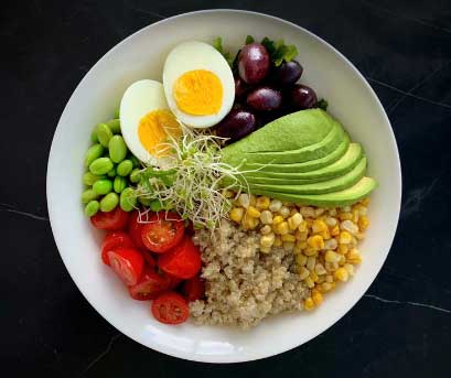Bowl of food containing quinoa, corn, avocado, black olives, a hard boiled egg, edamame beans, cherry tomatoes and sprouts