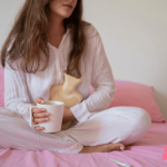 PMS and how naturopathy can help with it. Image of woman sat on bed cuddling a hot water bottle with a nice cup of tea in hand.