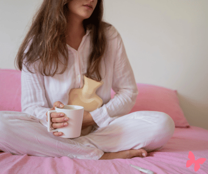 PMS and how naturopathy can help with it. Image of woman sat on bed cuddling a hot water bottle with a nice cup of tea in hand.