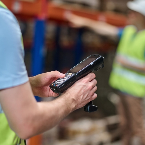 Barcode scanner. Hands of man in overalls holding black barcode scanner and colleague behind pointing to shelf with cargo at industrial warehouse