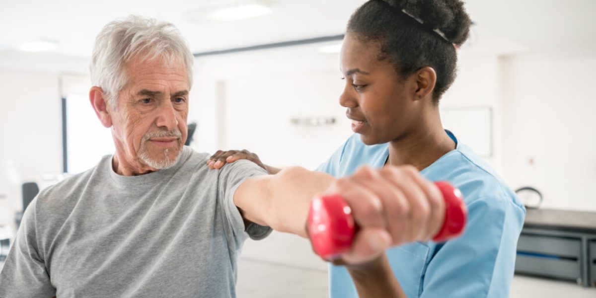 Physiotherapist correcting her senior patient with his shoulder posture as he lifts free weights both looking focused and smiling