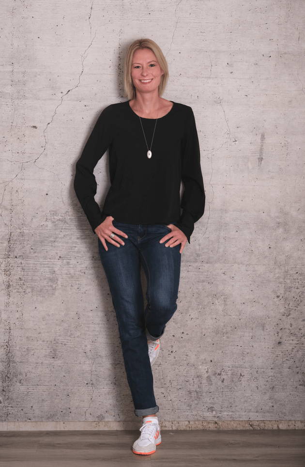 virtuelle-assistentin-melanie-fraunhofer melanie-fraunhofer-in-schwarzer-bluse-lehnt-laechelnd-an-grauer-beton-wand