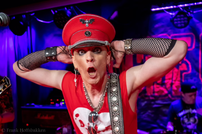 Chris Damien Doll A performer in a red military-style cap, fishnet sleeves, and studded accessories sings passionately on stage, illuminated by blue and purple stage lights.