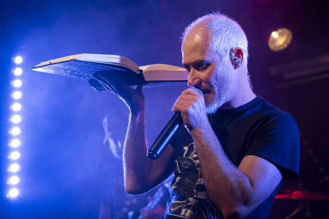 AMON-SETHIS (Live at Le Brin de Zinc, Barberaz, Chambéry, France, May 16, 2025) Julien Tournoud, singer of Amon Sethis, performing live. He's holding up a big old fashion book in a dramatic pose.