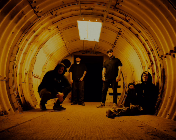 Mugshot band photo four men, one kneeling down with his hood up, two standing, one sitting on the ground with his hood up, all staring at the camera in a lit up tunnel.