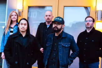 four males, one female staring ahead, on male has his hands in his pocket with a baseball hat, the other three men wearing black also hands in their pocket, female wearing a blue jean jackett