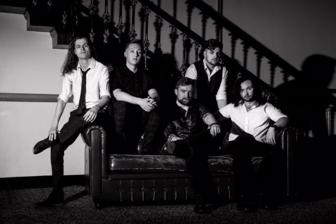 Skies Turn Black 2025 A black and white photo of the five members of the band Skies Turn Black. Two members are seated on a dark leather couch or bench, two are seated on the back of the couch, and one is seated on the armrest. A staircase with an ornate railing is visible in the background.
