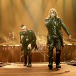 A full-shot photo of Matt Walst and Adam Gontier of Three Days Grace performing live at Rogers Place in Edmonton, Canada. Matt Walst, on the left, is wearing a black jacket and pants, with his left hand on his knee, leaning into a microphone. Adam Gontier, on the right, is wearing a long black leather coat and dark pants, singing into a microphone. Both are illuminated by warm stage lights, with a drum kit visible in the background.