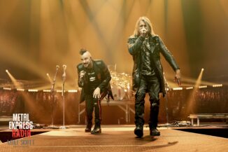 A full-shot photo of Matt Walst and Adam Gontier of Three Days Grace performing live at Rogers Place in Edmonton, Canada. Matt Walst, on the left, is wearing a black jacket and pants, with his left hand on his knee, leaning into a microphone. Adam Gontier, on the right, is wearing a long black leather coat and dark pants, singing into a microphone. Both are illuminated by warm stage lights, with a drum kit visible in the background.