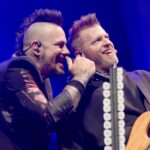 A close-up photo of Matt Walst and Brad Walst of Three Days Grace performing live. Matt Walst, on the left, with a mohawk, is leaning in towards Brad Walst, on the right, who is holding a bass guitar. Both are smiling broadly and appear to be interacting playfully during the concert. Matt is holding a microphone near Brad's ear. The background is dark, focusing attention on the two musicians.
