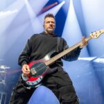A dynamic, low-angle photo of Brad Walst, the bassist for Three Days Grace, performing live on stage. He is looking upwards and to the right, intensely focused on his playing. Brad is wearing a dark hoodie and dark pants, and he is holding a reddish-black bass guitar with a light-colored neck. Bright white and blue stage lights illuminate the scene from behind and above him, creating a dramatic atmosphere, with a drum set partially visible in the background on the left.