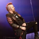 A dynamic, mid-shot photo of Matt Walst, the co-lead vocalist of Three Days Grace, performing on stage. He is captured mid-action, leaning back with one leg bent and foot planted firmly, and his left arm extended, holding a microphone. Matt has a prominent red mohawk and is wearing a black t-shirt under a black jacket with red distressed or patterned accents on the sleeves and body. He is also wearing ripped black pants and dark boots. The stage is dimly lit with purplish-blue light in the background, creating a dramatic and energetic atmosphere.