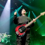 A wide-angle, low-shot photo of Brad Walst, the bassist for Three Days Grace, on stage during a performance. He is looking upwards and to the right, with a smile on his face, and his left hand is raised with his middle finger pointing up. He is holding a reddish-black bass guitar. Brad is wearing a dark hoodie and dark pants. The stage is dramatically lit with multiple bright green and white spotlights shining down from above, creating a strong contrast. A drum kit is visible in the background to the left.