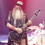 A mid-shot photo of Barry Stock, the guitarist for Three Days Grace, performing live on stage. He is looking directly at the camera with a serious expression, playing a black electric guitar with a distinctive body shape and white accents. Barry has a very long, full gray beard and is wearing a baseball cap, a dark t-shirt, and a long black leather coat. His tattooed arms are visible, and he has a red wristband on his right wrist. The stage is lit with red and pink lights, and a drummer is visible in the blurry background to the right.