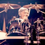 A close-up, mid-shot photo of Jon Larsen, the drummer for Volbeat, behind his drum kit on stage. He is smiling and looking towards the right, with a drumstick in his right hand. Jon is wearing a camouflage patterned cap and a light-colored t-shirt. Various cymbals and drum components, including a tom drum directly in front of him, are visible. The stage lighting behind him appears dark.