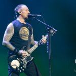 A mid-shot photo of Michael Poulsen, the lead vocalist and guitarist of Volbeat, performing live on stage. He is singing into a microphone while playing a dark-colored electric guitar with white accents. Michael is wearing a black sleeveless t-shirt with a gold symbol on the front, black pants, and has visible tattoos on both arms. He has short hair and a goatee. The stage is dimly lit with blue and green lighting, creating a focused and intense atmosphere.