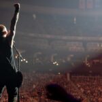 A wide-angle photo of Michael Poulsen of Volbeat from behind, facing a massive crowd in a large arena. He is standing on stage, holding his guitar, with his right arm raised in the air with a clenched fist, engaging with the audience. The arena seating is packed with concert-goers, visible as a sea of faces and lights in the background. Bright spotlights illuminate the stage and part of the crowd from above, creating a grand and energetic concert atmosphere.