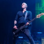 A dynamic, low-angle photo of Kaspar Boye Larsen, the bassist for Volbeat, performing live on stage. He is looking upwards with a focused expression, deeply engaged in playing his black bass guitar. Kaspar is wearing a dark, long-sleeved collared shirt with a pattern, dark pants, and glasses. Bright blue and green spotlights illuminate him from behind and above, creating dramatic light rays on the dark stage. Part of his amplifier rig is visible in the lower right corner.