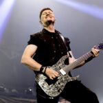 A dynamic, low-angle photo of Flemming C. Lund, the lead guitarist for Volbeat, performing live on stage. He is looking upwards with a joyful expression, eyes closed, playing a black electric guitar with a white splatter or marble-like pattern. Flemming is wearing a black collared shirt with the sleeves rolled up and dark pants. Bright white spotlights shine down from above, illuminating him against a smoky, dark background.