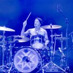 A wide-angle, eye-level photo of Stephen Kluesener, the drummer for Wage War, intensely focused on his drum kit during a live performance. He is shirtless, revealing tattooed arms and chest, and is wearing a dark beanie. He holds a drumstick aloft in his right hand, poised to strike. The drums are a dark metallic color, with the bass drum featuring a prominent white, geometric star-like logo. The stage is bathed in bright, vibrant blue light from above, creating a cool and energetic atmosphere.