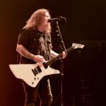 A mid-shot photo of Cody Quistad, the rhythm guitarist and clean vocalist for Wage War, performing live on stage. He is looking up and singing into a microphone while playing a white electric guitar with a distinct angular body. Cody has long, wavy brown hair that falls over his shoulders and is wearing a dark, short-sleeved collared shirt over a black t-shirt, with numerous visible tattoos on his arms. The stage is bathed in warm, orange-red lighting from vertical light bars in the background, creating a powerful and energetic atmosphere.
