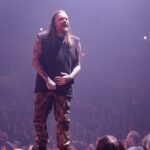 A wide-angle shot of Briton Bond, the lead vocalist of Wage War, standing on stage and looking out at the audience. He has long, dark brown hair and is wearing a black sleeveless t-shirt, revealing tattooed arms, and camouflage cargo pants. His hands are clasped in front of him. The stage is bathed in a pinkish-purple light, and the heads of the crowd are visible in the blurry foreground, emphasizing the scale of the concert.